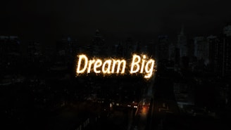A nighttime cityscape with tall buildings in the background. The words 'Dream Big' are illuminated in bright, glowing letters across the center of the image, resembling a light display or sparkler effect.