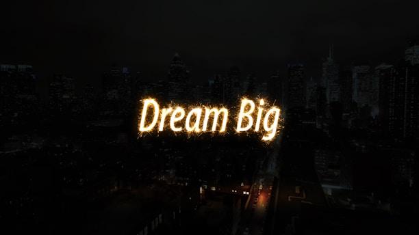 A nighttime cityscape with tall buildings in the background. The words 'Dream Big' are illuminated in bright, glowing letters across the center of the image, resembling a light display or sparkler effect.