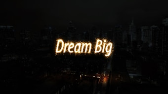 A nighttime cityscape with tall buildings in the background. The words 'Dream Big' are illuminated in bright, glowing letters across the center of the image, resembling a light display or sparkler effect.