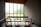 A minimalist interior space with wooden chairs arranged facing a large window panel. The window has frosted glass allowing diffused natural light to enter. The walls are textured and the ceiling is wooden, adding warmth to the serene atmosphere. The room has a quiet and contemplative mood.