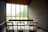 A minimalist interior space with wooden chairs arranged facing a large window panel. The window has frosted glass allowing diffused natural light to enter. The walls are textured and the ceiling is wooden, adding warmth to the serene atmosphere. The room has a quiet and contemplative mood.