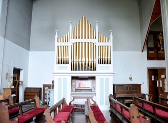 An interior shot of a church featuring an ornate pipe organ with gold and white pipes. Wooden pews with red cushions are arranged facing the organ, and there is other wooden furniture, including chairs and a small table with vases, visible in the space.