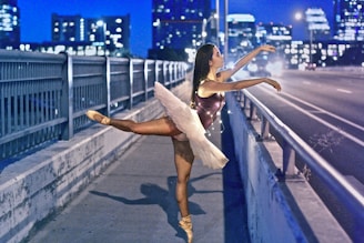 A ballerina in a white tutu and a dark leotard is gracefully posing on a bridge at night. The city skyline and lit-up buildings are visible in the background, creating a contrasting urban setting. The bridge is adorned with streetlights, and the road is empty, emphasizing the serene yet dynamic atmosphere.