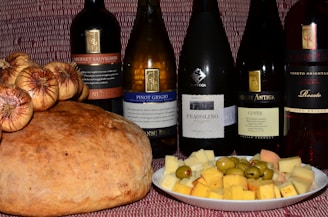 A variety of wine bottles are displayed alongside a large loaf of bread topped with dried figs and a plate of assorted cheese cubes and olives. The background has a textured red pattern.