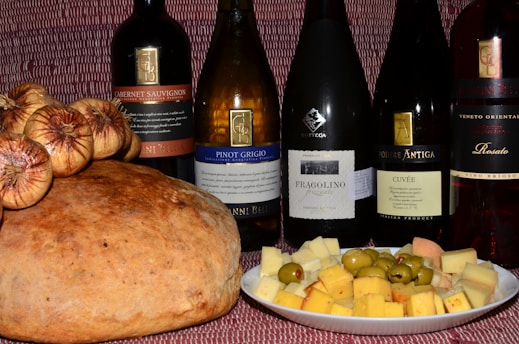 A variety of wine bottles are displayed alongside a large loaf of bread topped with dried figs and a plate of assorted cheese cubes and olives. The background has a textured red pattern.