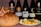 A variety of wine bottles are displayed alongside a large loaf of bread topped with dried figs and a plate of assorted cheese cubes and olives. The background has a textured red pattern.