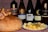 A variety of wine bottles are displayed alongside a large loaf of bread topped with dried figs and a plate of assorted cheese cubes and olives. The background has a textured red pattern.