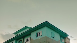 A pastel-colored house with teal walls and a dark teal roof is depicted against a soft, overcast sky. The house features multiple windows with shutters, some open, on its upper stories.