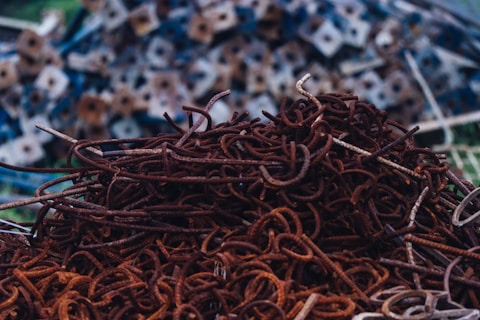 A pile of rusty metal rods is tangled together, creating a chaotic arrangement of twisted shapes. In the background, there are blurred, geometric metal pieces stacked on top of each other, adding depth to the scene.