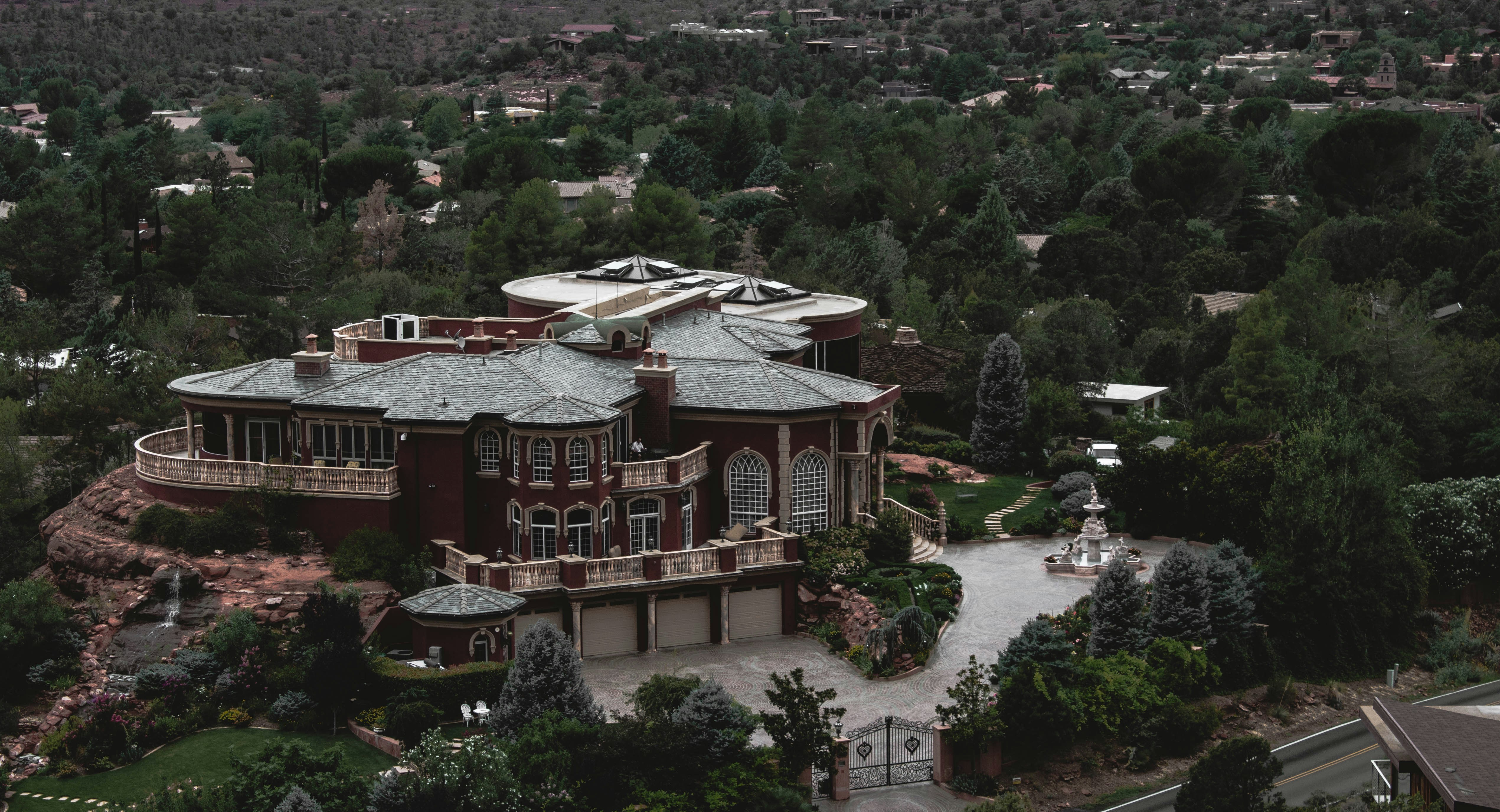 A large, luxurious mansion surrounded by lush greenery and trees, featuring a grand architectural design with multiple arched windows, a circular driveway, and a water fountain in front. The building is set against a backdrop of a forested area with many trees extending into the distance.