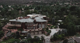 A large, luxurious mansion surrounded by lush greenery and trees, featuring a grand architectural design with multiple arched windows, a circular driveway, and a water fountain in front. The building is set against a backdrop of a forested area with many trees extending into the distance.