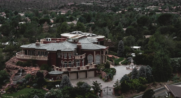 A large, luxurious mansion surrounded by lush greenery and trees, featuring a grand architectural design with multiple arched windows, a circular driveway, and a water fountain in front. The building is set against a backdrop of a forested area with many trees extending into the distance.
