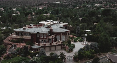 A large, luxurious mansion surrounded by lush greenery and trees, featuring a grand architectural design with multiple arched windows, a circular driveway, and a water fountain in front. The building is set against a backdrop of a forested area with many trees extending into the distance.