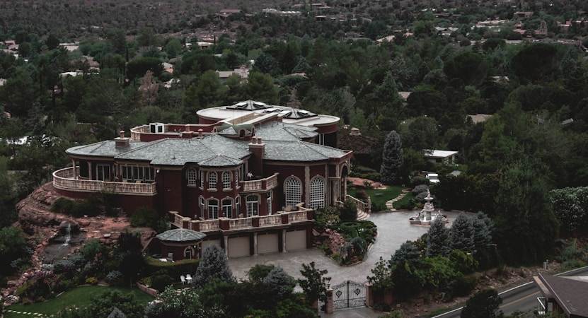 A large, luxurious mansion surrounded by lush greenery and trees, featuring a grand architectural design with multiple arched windows, a circular driveway, and a water fountain in front. The building is set against a backdrop of a forested area with many trees extending into the distance.