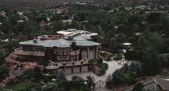 A large, luxurious mansion surrounded by lush greenery and trees, featuring a grand architectural design with multiple arched windows, a circular driveway, and a water fountain in front. The building is set against a backdrop of a forested area with many trees extending into the distance.