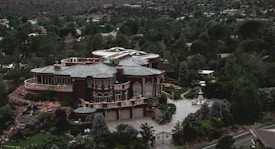 A large, luxurious mansion surrounded by lush greenery and trees, featuring a grand architectural design with multiple arched windows, a circular driveway, and a water fountain in front. The building is set against a backdrop of a forested area with many trees extending into the distance.