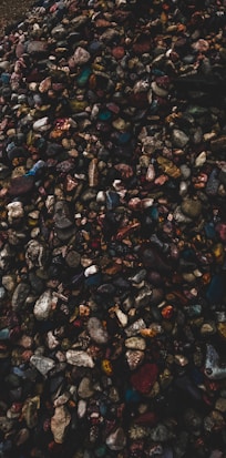A pile of assorted pebbles and stones in various sizes and colors, including grays, blues, reds, and browns. The image has a textured and uneven surface with some darker areas creating a contrast.
