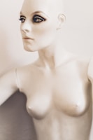 A mannequin with striking makeup, featuring pronounced eyeliner and mascara on the eyes, creating a dramatic look. The mannequin has a pale, smooth surface and is positioned against a plain background.