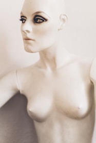 A mannequin with striking makeup, featuring pronounced eyeliner and mascara on the eyes, creating a dramatic look. The mannequin has a pale, smooth surface and is positioned against a plain background.