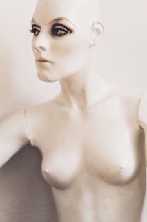 A mannequin with striking makeup, featuring pronounced eyeliner and mascara on the eyes, creating a dramatic look. The mannequin has a pale, smooth surface and is positioned against a plain background.