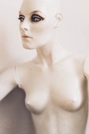 A mannequin with striking makeup, featuring pronounced eyeliner and mascara on the eyes, creating a dramatic look. The mannequin has a pale, smooth surface and is positioned against a plain background.