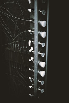 A section of an old electrical panel is visible, featuring a series of switches or knobs aligned vertically. The panel is surrounded by numerous thin wires that appear to be tangled. The image gives a vintage, industrial feel due to the black and white color scheme.