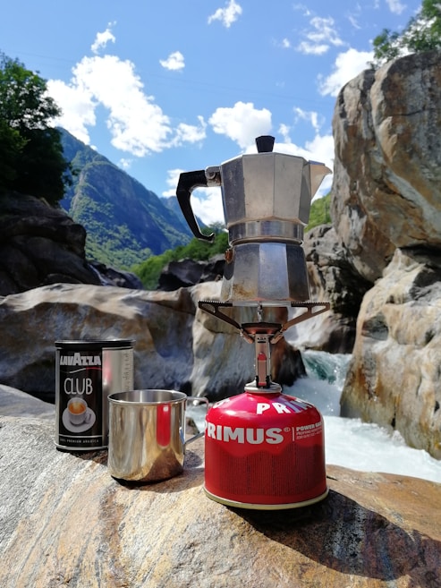 A Moka pot is placed on a portable gas stove atop a rock in a nature setting, accompanied by a can of Lavazza coffee and a metal cup. The background features rocky terrain, a waterfall, and lush green mountains under a partly cloudy blue sky.