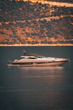 A luxurious yacht is floating on calm waters with a backdrop of a steep, wooded hillside. The light creates a golden reflection on the water.