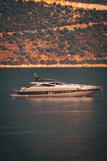 A luxurious yacht is floating on calm waters with a backdrop of a steep, wooded hillside. The light creates a golden reflection on the water.