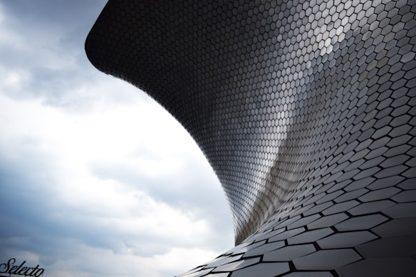 A sleek, modern structure with a curving design covered in hexagonal tiles against a cloudy sky backdrop. The building&rsquo;s surface reflects light, creating a dynamic visual effect.