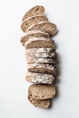Several slices of brown, whole-grain bread arranged in a vertical stack on a white surface. The bread appears to have a crusty texture with a dusting of flour on the top.