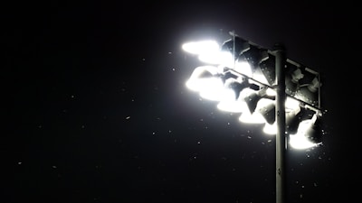 A set of bright stadium floodlights illuminating the surrounding darkness. Small particles or insects are visible in the light beam, adding a dynamic element to the scene.
