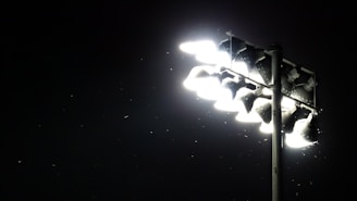 A set of bright stadium floodlights illuminating the surrounding darkness. Small particles or insects are visible in the light beam, adding a dynamic element to the scene.