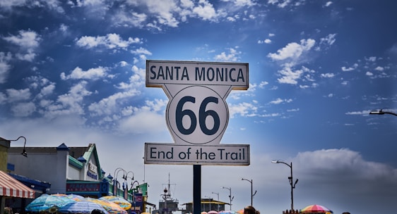 A prominent Route 66 sign stands with the text 'Santa Monica, End of the Trail'. The background features a partly cloudy sky. Below, colorful umbrellas line the street, suggesting a market or outdoor commercial area with various shops and buildings.