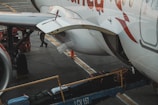 The image captures a close-up of an airplane on the tarmac with luggage being loaded onto it. A conveyor belt transports suitcases into the cargo hold. Part of the aircraft's fuselage and wing are visible, with a person in the background attending to the loading process.