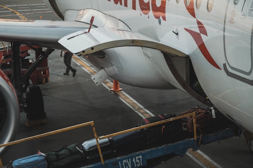 The image captures a close-up of an airplane on the tarmac with luggage being loaded onto it. A conveyor belt transports suitcases into the cargo hold. Part of the aircraft's fuselage and wing are visible, with a person in the background attending to the loading process.