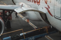 The image captures a close-up of an airplane on the tarmac with luggage being loaded onto it. A conveyor belt transports suitcases into the cargo hold. Part of the aircraft's fuselage and wing are visible, with a person in the background attending to the loading process.