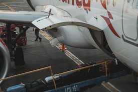 The image captures a close-up of an airplane on the tarmac with luggage being loaded onto it. A conveyor belt transports suitcases into the cargo hold. Part of the aircraft's fuselage and wing are visible, with a person in the background attending to the loading process.