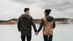 couple holding hand front of body of calm water with mountain distance