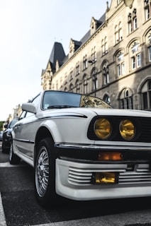 A vintage white car parked on a street with a historic stone building in the background. The car features distinctive yellow headlights and stylish alloy wheels, creating a classic, nostalgic vibe. The architecture of the building showcases arched windows and intricate stonework, reflecting a traditional European style.