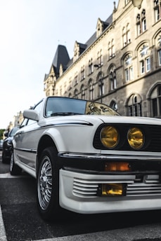 A vintage white car parked on a street with a historic stone building in the background. The car features distinctive yellow headlights and stylish alloy wheels, creating a classic, nostalgic vibe. The architecture of the building showcases arched windows and intricate stonework, reflecting a traditional European style.