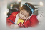 A young girl is focused on writing at a table, holding a blue pencil in her right hand while resting her head on her left hand. She wears a red jacket over a yellow sweater and has a blue headband with floral decorations in her hair. Next to her is a white plush toy with pink ears and nose.