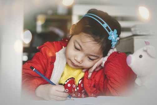 A young girl is focused on writing at a table, holding a blue pencil in her right hand while resting her head on her left hand. She wears a red jacket over a yellow sweater and has a blue headband with floral decorations in her hair. Next to her is a white plush toy with pink ears and nose.