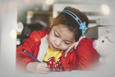 A young girl is focused on writing at a table, holding a blue pencil in her right hand while resting her head on her left hand. She wears a red jacket over a yellow sweater and has a blue headband with floral decorations in her hair. Next to her is a white plush toy with pink ears and nose.