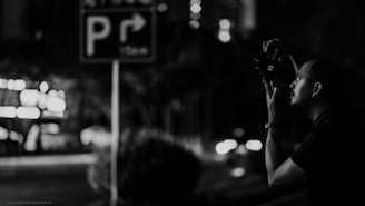 Black and white photo of a photographer capturing a candid street moment with soft purple and charcoal tones in the background.