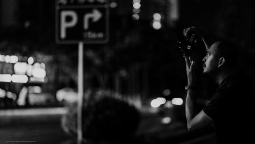 A black and white photograph captures a person holding a camera, presumably taking a picture at night. In the background, there is a sign with a parking symbol and an arrow, and blurred lights suggest an urban street setting with cars passing by.