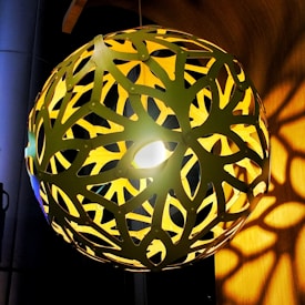 A spherical lamp with intricate cut-out patterns casts shadows on the surrounding surfaces. The design resembles overlapping leaves, and the light source from within illuminates the space with a warm glow.