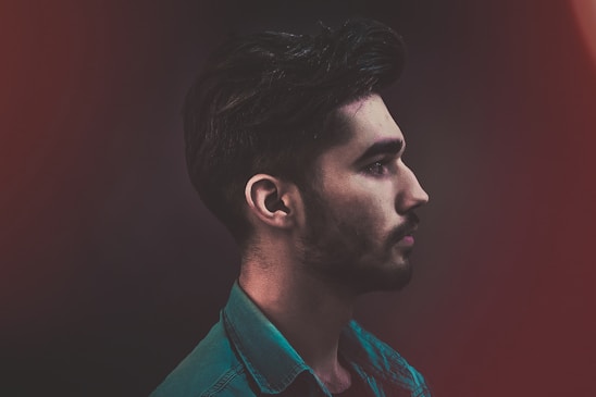 A profile view of a young man with a beard, wearing a teal denim shirt against a dark, blurred background with subtle red hues.