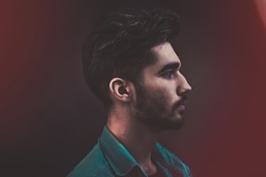 A profile view of a young man with a beard, wearing a teal denim shirt against a dark, blurred background with subtle red hues.