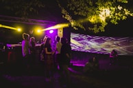 A group of people gathered outdoors at night, illuminated by vibrant yellow and purple lights. In the background, a DJ stands at a booth, and a large screen displays abstract patterns. The area is surrounded by trees with bright lights shining through the leaves, creating a festive atmosphere.